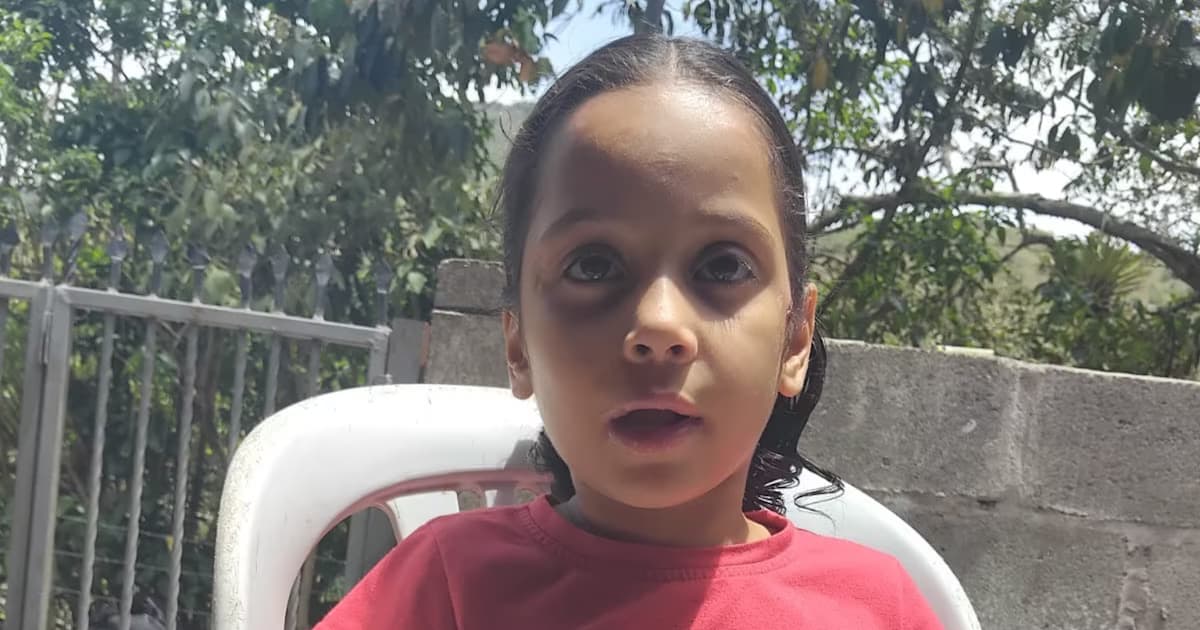 Retrato de una niña de pelo oscuro recogido y camiseta roja, sentada en una silla plástica blanca en un patio con árboles y un muro de bloques al fondo
