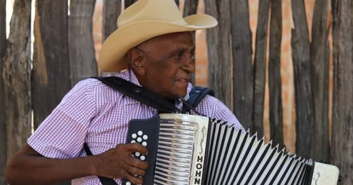 Náfer Durán, con sombrero claro, toca el acordeón frente a una cerca de madera