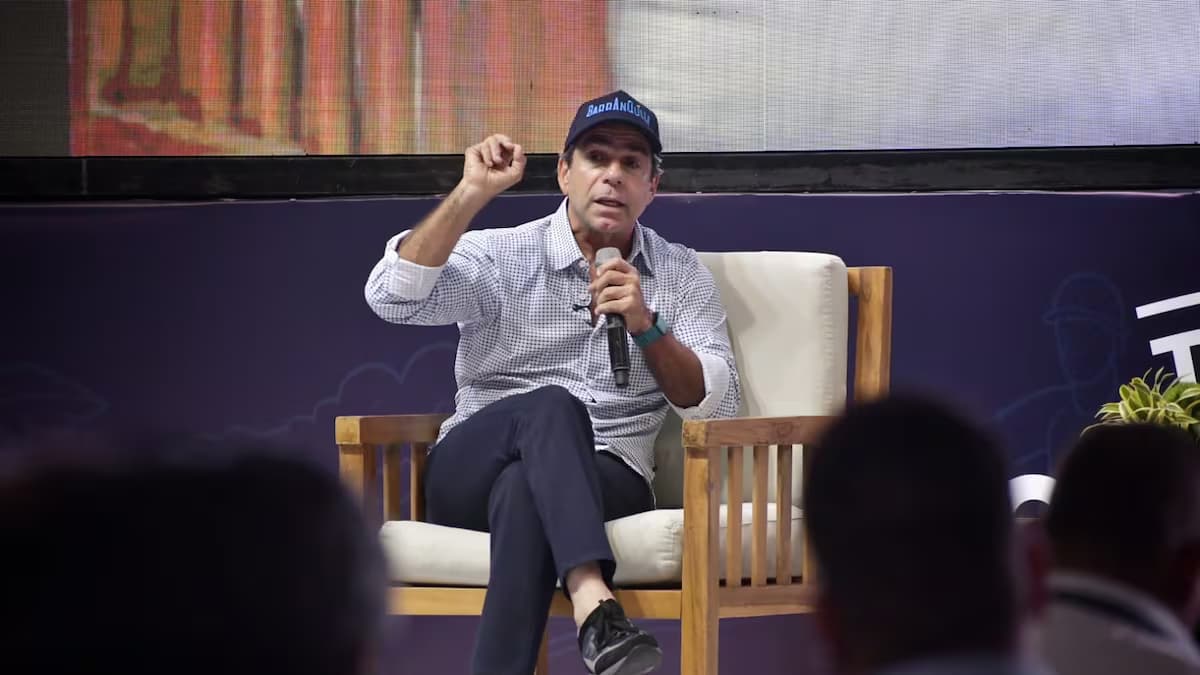 El alcalde de Barranquilla, Alejandro Char, con gorra azul y micrófono en mano, sentado en un sillón durante un acto público