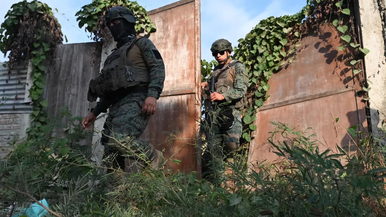 Dos militares con uniforme de camuflaje y fusiles avanzan entre la maleza junto a un portón metálico oxidado