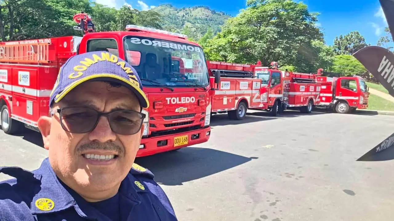 Un bombero con gorra de comandante y uniforme azul posa frente a una fila de máquinas de bomberos rojas estacionadas en una vía