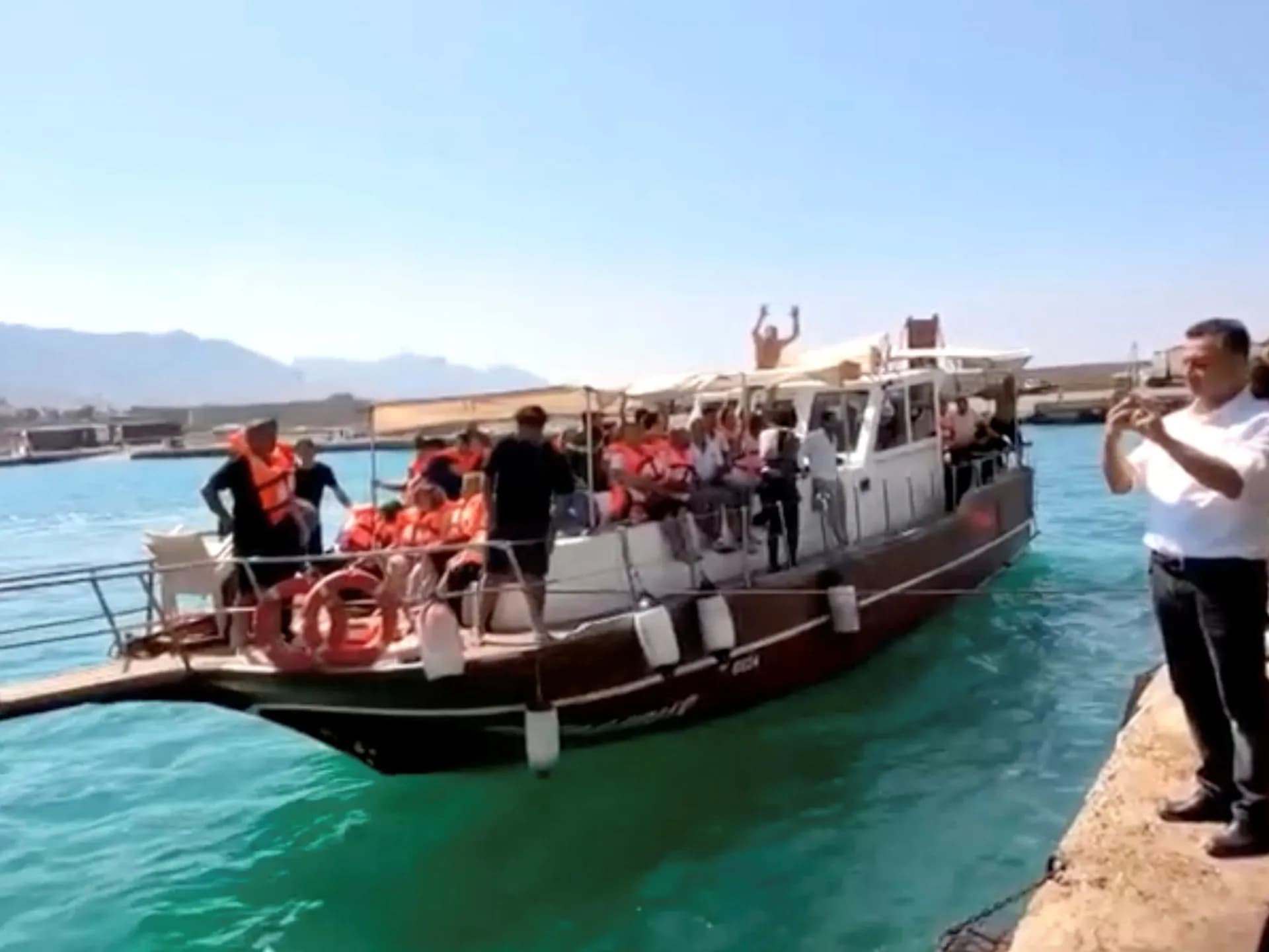 Una embarcación de rescate llega al muelle cargada de personas con chalecos salvavidas naranjas mientras un hombre las graba desde el malecón
