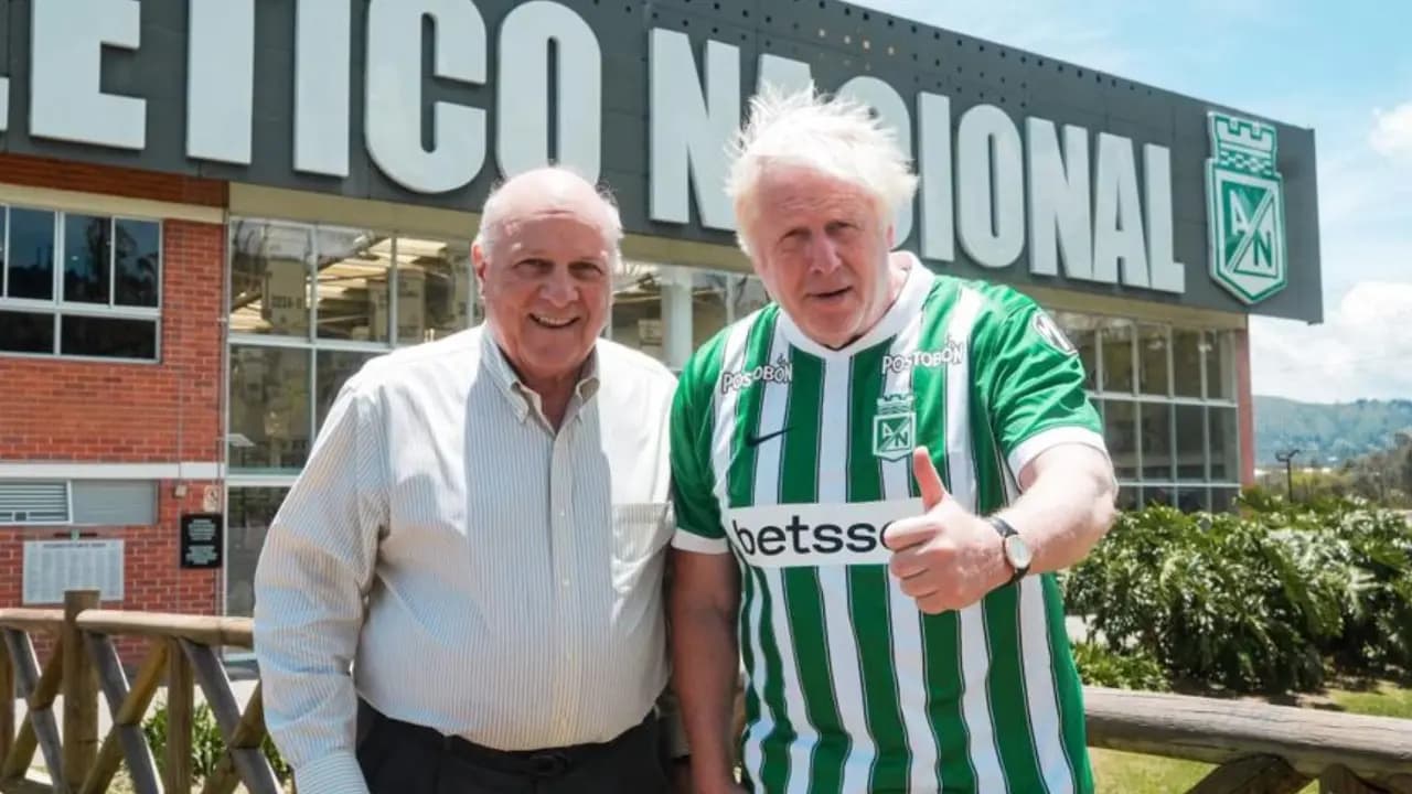 Boris Johnson, con la camiseta verdiblanca de Atlético Nacional, posa junto a un directivo del club frente al letrero de la sede de Guarne