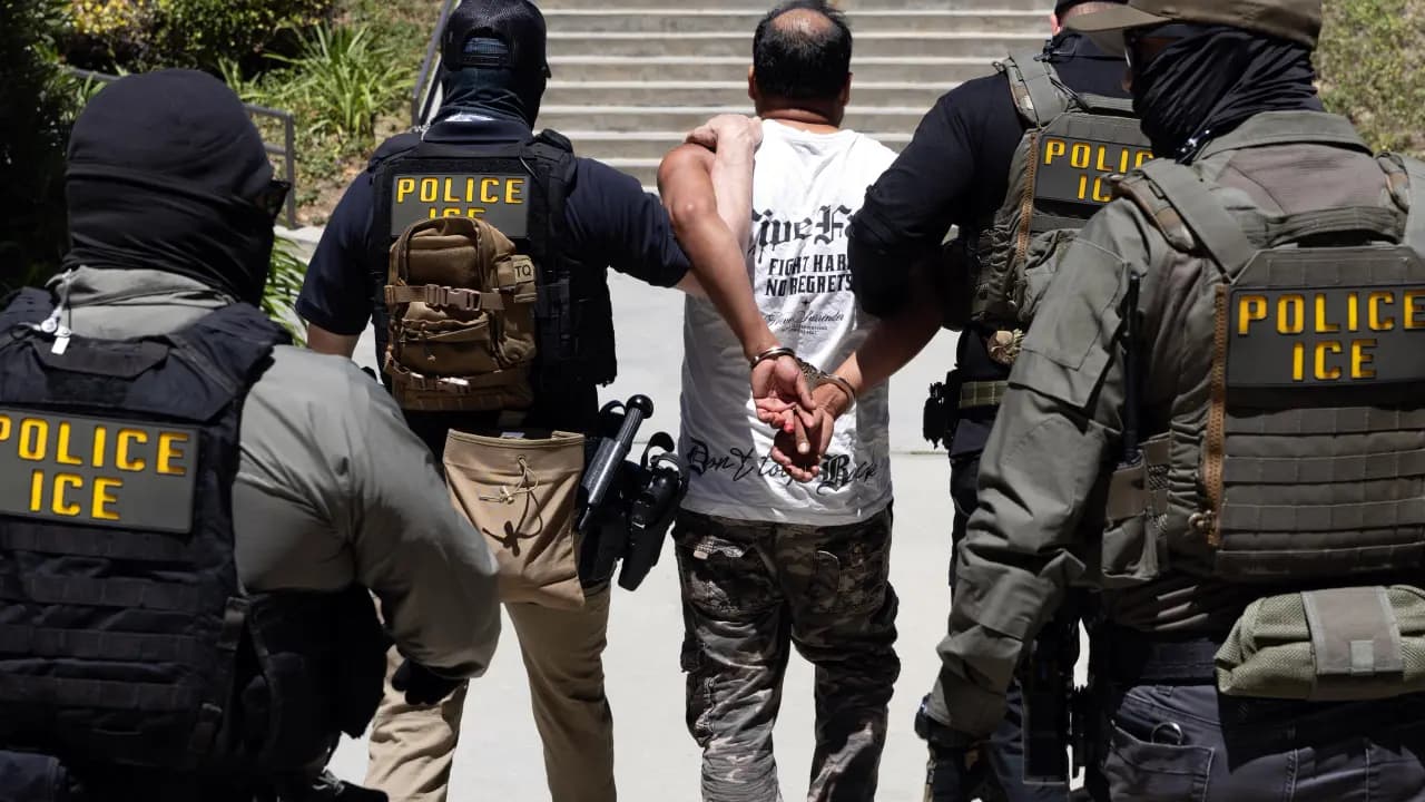 Agentes con chalecos que dicen Police ICE conducen esposado a un hombre de camiseta blanca por un andén