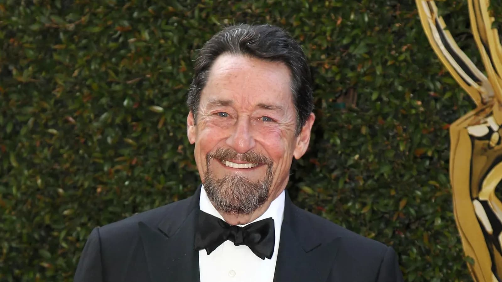 Peter Cullen sonríe de esmoquin y corbatín negro ante un fondo de vegetación
