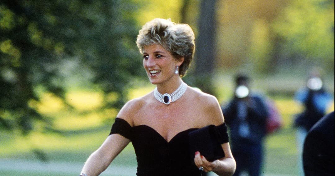 La princesa Diana sonríe con un vestido negro de hombros descubiertos y un collar de perlas con una piedra oscura, en un jardín