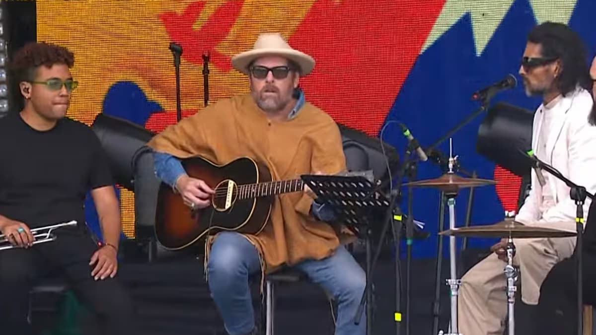 Un músico con sombrero y ruana toca la guitarra sentado en el escenario del concierto junto a otros intérpretes