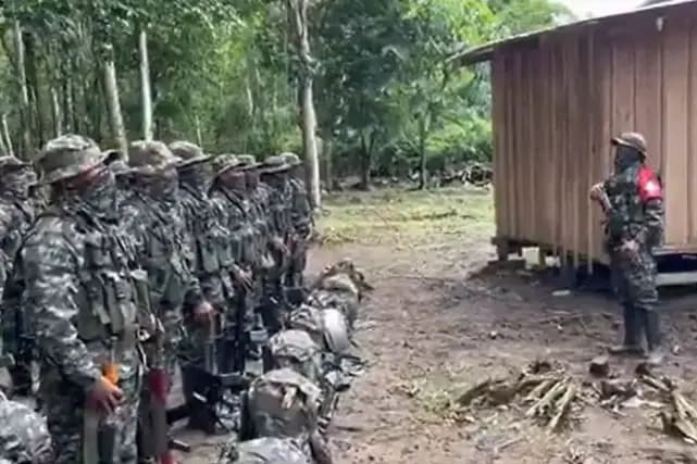 Una fila de hombres uniformados de camuflado y con fusiles permanece formada frente a una construcción de madera, en un claro rodeado de monte