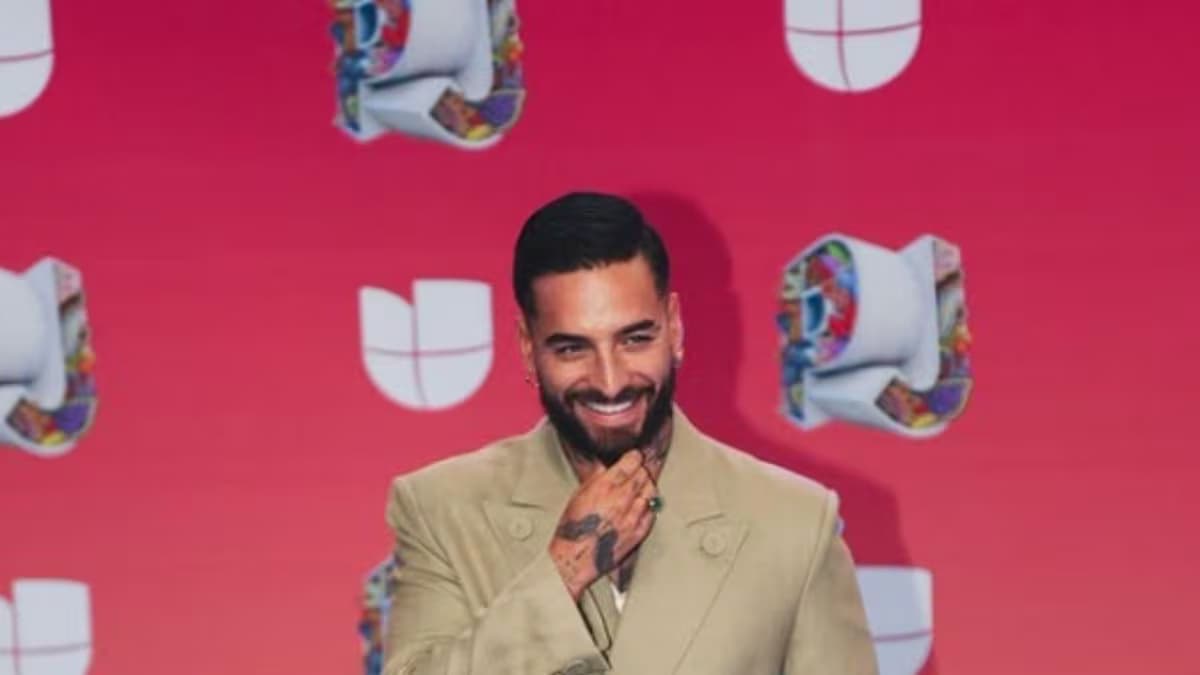 Maluma sonríe con traje beige frente a un fondo rojo con logos de Univision