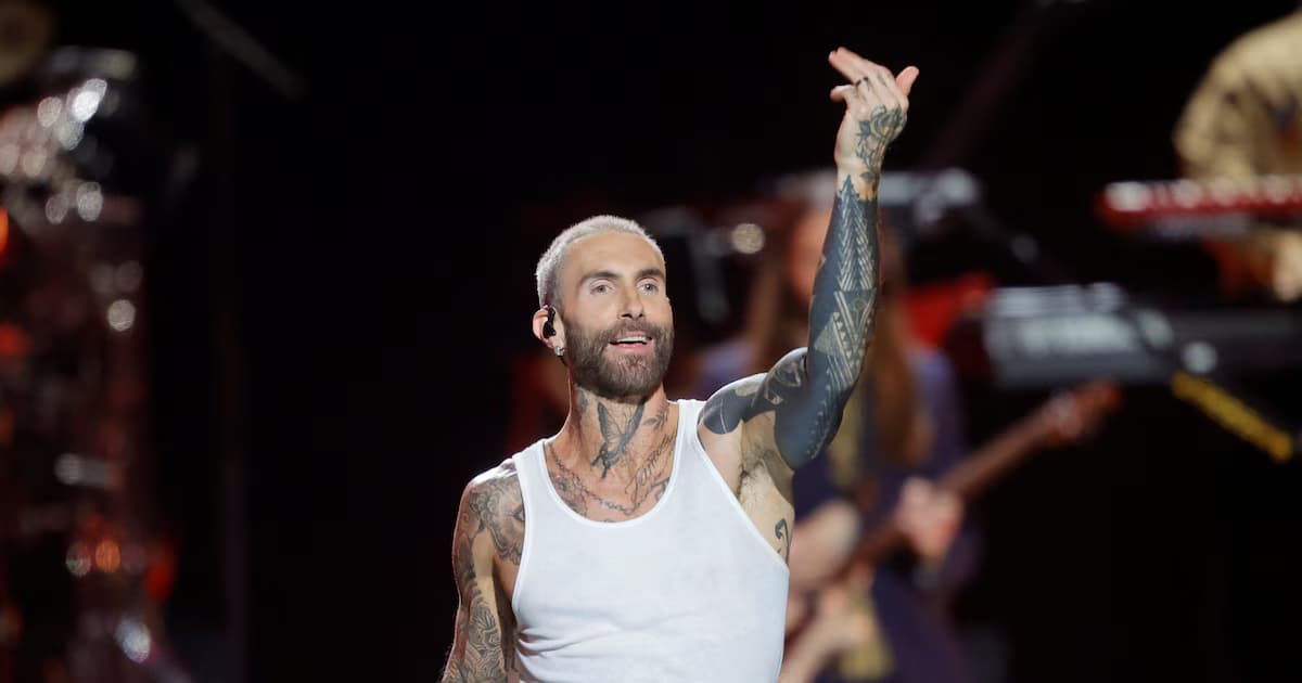 Adam Levine canta sobre el escenario con una camiseta blanca sin mangas y el brazo derecho levantado