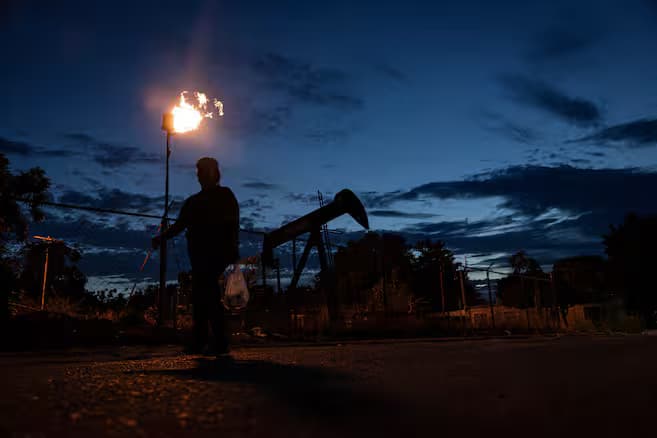 Silueta de un hombre que camina al anochecer junto a un mechero encendido y un balancín de extracción petrolera