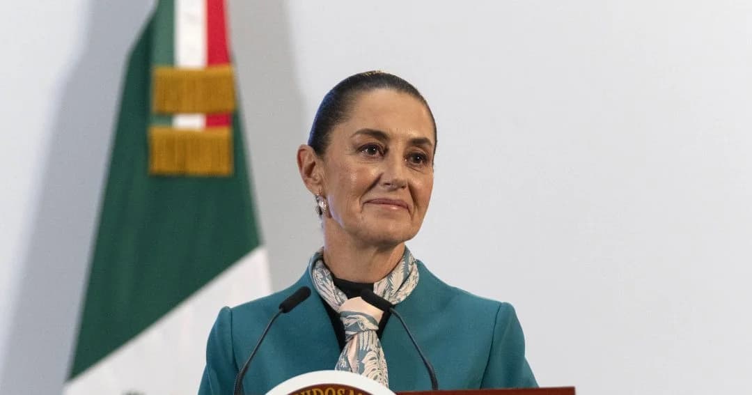 Claudia Sheinbaum, de saco verde azulado y pañuelo estampado, habla en un atril junto a la bandera de México