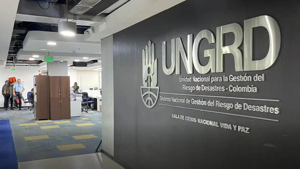 Muro gris de una oficina con las letras metálicas UNGRD y el nombre completo de la Unidad Nacional para la Gestión del Riesgo de Desastres, con escritorios y empleados al fondo