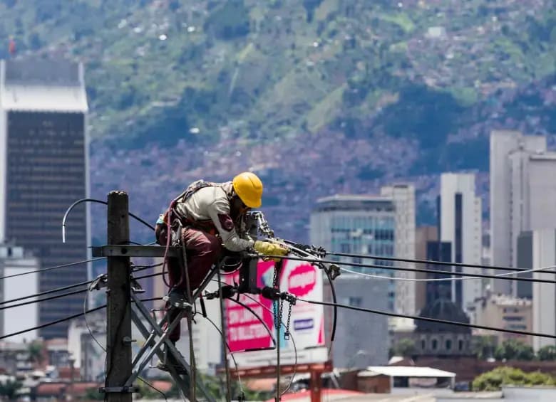 Un operario con casco amarillo trabaja sobre un poste de energía con la ciudad de Medellín al fondo