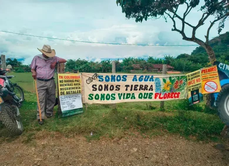 Un campesino con sombrero y bastón se apoya en un poste junto a pancartas pintadas a mano que dicen «Somos agua, somos tierra, somos vida que florece» al borde de una carretera destapada