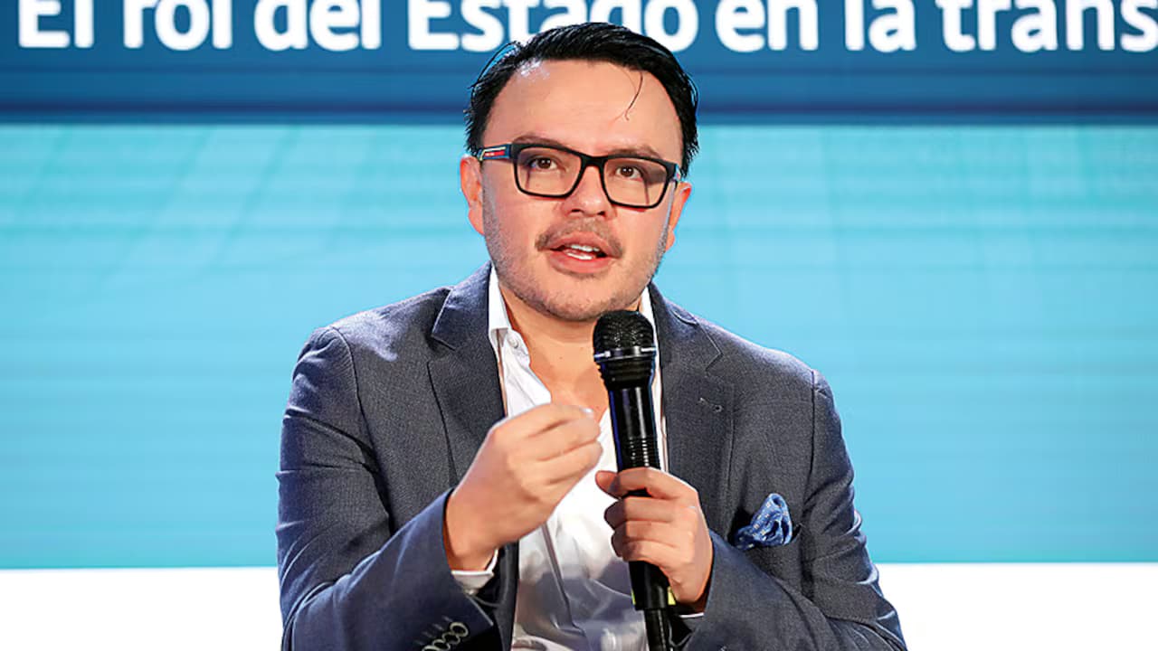 Juan Felipe Harman, de saco gris y gafas oscuras de pasta, habla con un micrófono en la mano durante un foro, sentado frente a un telón azul