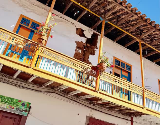 Balcón de madera amarilla de una casa colonial con una grieta profunda que atraviesa el muro blanco bajo el alero de tejas