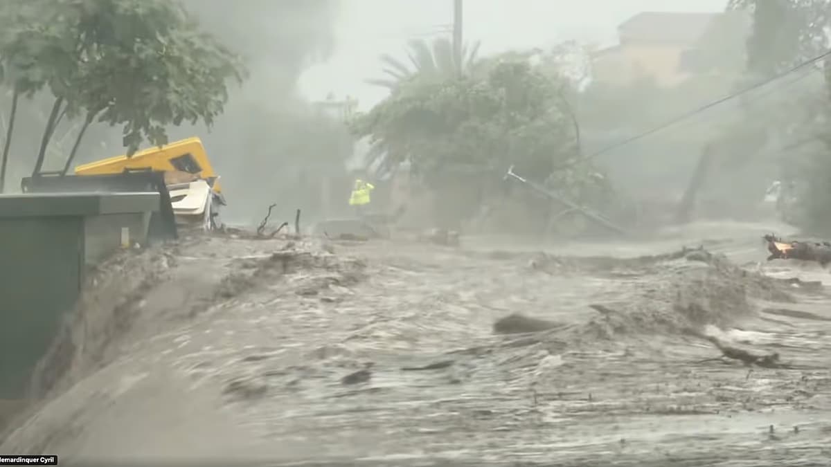 Una calle convertida en un torrente de agua y lodo bajo la lluvia, con un rescatista con chaleco amarillo al fondo, en el sur de Francia