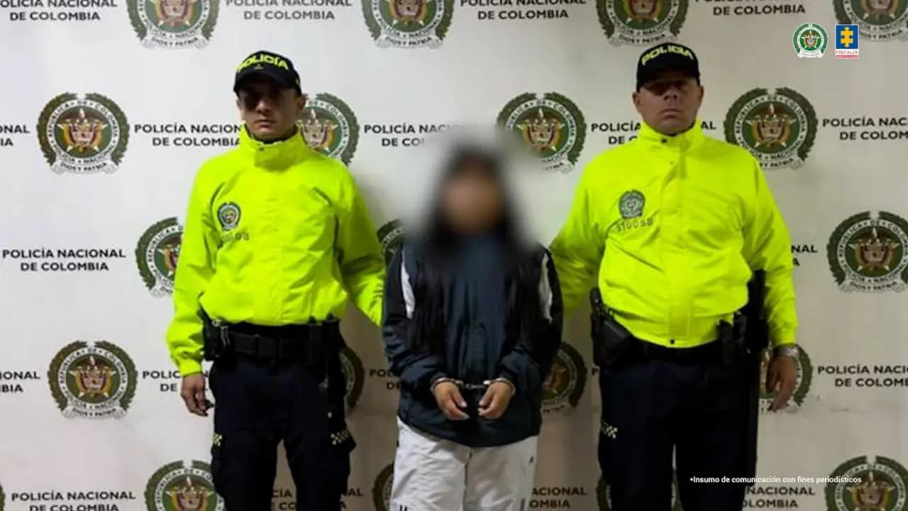 Dos policías custodian a la mujer capturada, con el rostro difuminado, frente a un pendón de la Policía Nacional