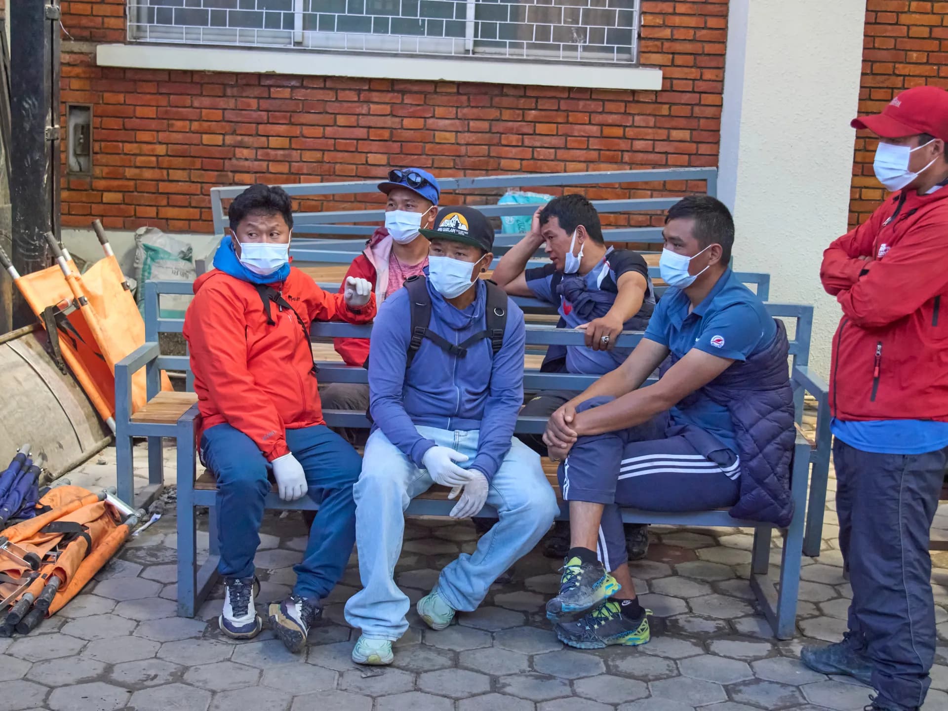 Rescatistas y guías nepalíes con tapabocas esperan sentados en una banca junto a camillas plegadas