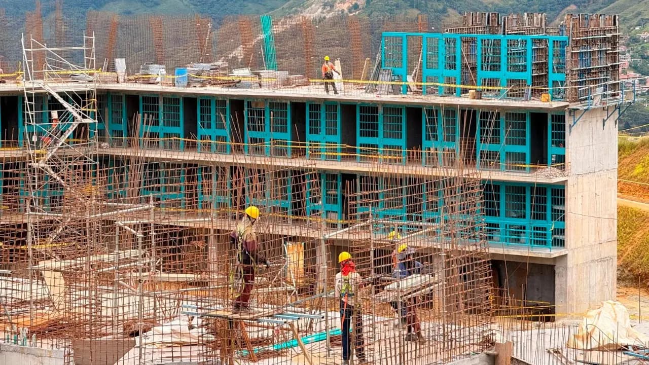 Obreros con casco trabajan entre andamios y varillas en los pabellones en construcción de la cárcel, con las montañas de fondo