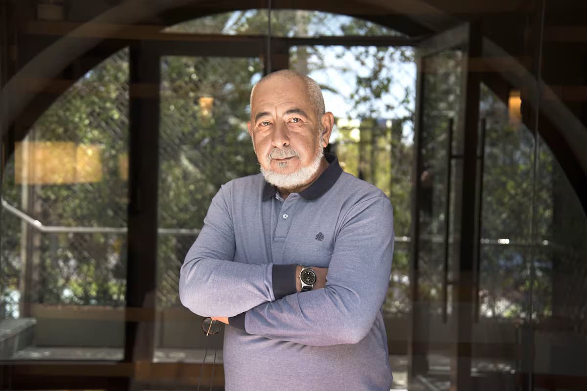 El escritor cubano Leonardo Padura, de barba blanca y suéter azul, posa con los brazos cruzados bajo un arco con puertas de vidrio