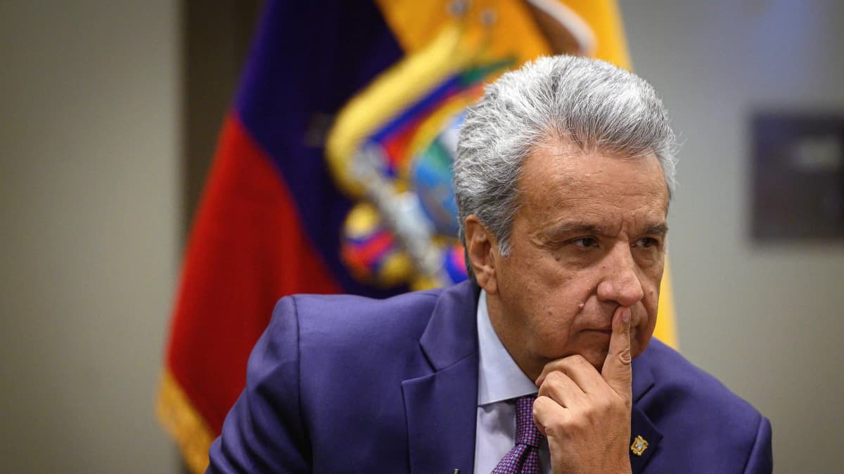 Lenín Moreno, con traje azul, se lleva un dedo a los labios sentado frente a la bandera de Ecuador