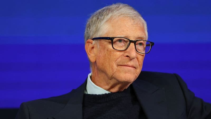 Bill Gates, con gafas de pasta y saco oscuro, escucha sentado en un escenario con fondo azul
