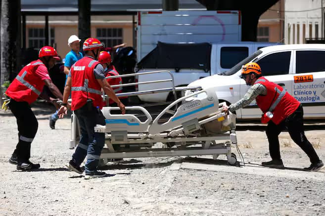 Cuatro socorristas con cascos y chalecos rojos empujan una camilla hospitalaria por un terreno destapado frente a un edificio de salud