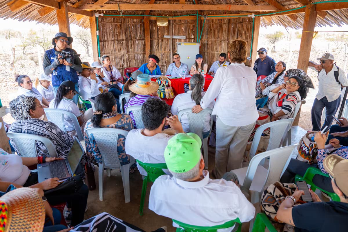 Decenas de personas sentadas en sillas plásticas bajo una enramada de palma en La Guajira escuchan a funcionarios sentados tras una mesa con mantel rojo, en una reunión con comunidades wayuu