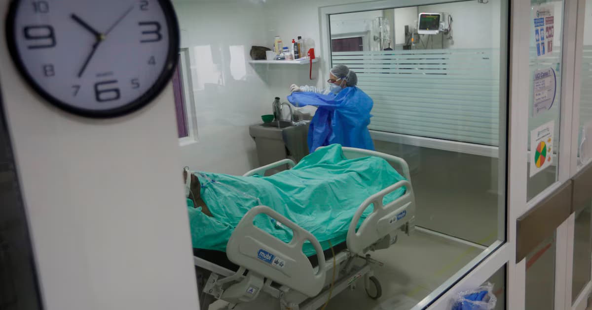 Vista a través de un vidrio de una habitación hospitalaria con una cama vacía tendida con sábanas verdes y un profesional de la salud con bata azul y tapabocas de espaldas