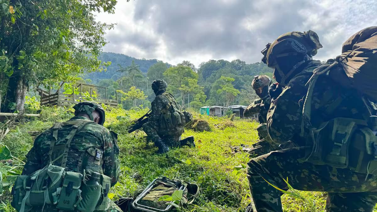 Soldados con uniforme camuflado, casco y fusil, agachados en un potrero rodeado de montaña