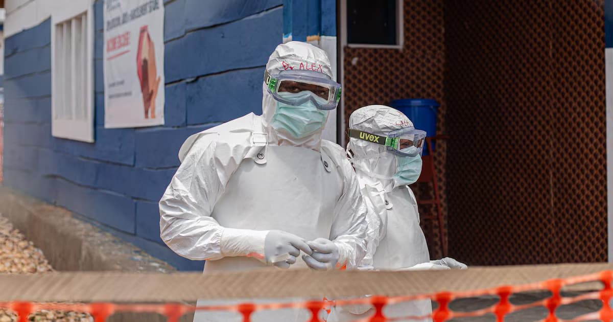 Dos trabajadores sanitarios con trajes de protección blancos y gafas frente a un centro de tratamiento