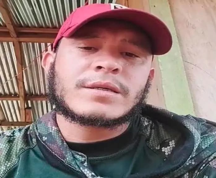 Retrato de cerca de alias Leito, con gorra roja, barba y chaqueta camuflada, bajo un techo de zinc