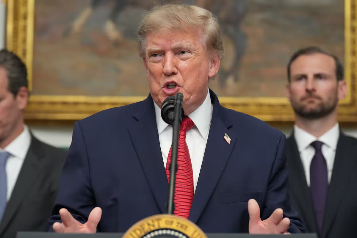El presidente Donald Trump habla ante un micrófono en un atril con el sello presidencial, con las manos abiertas y dos hombres de traje detrás