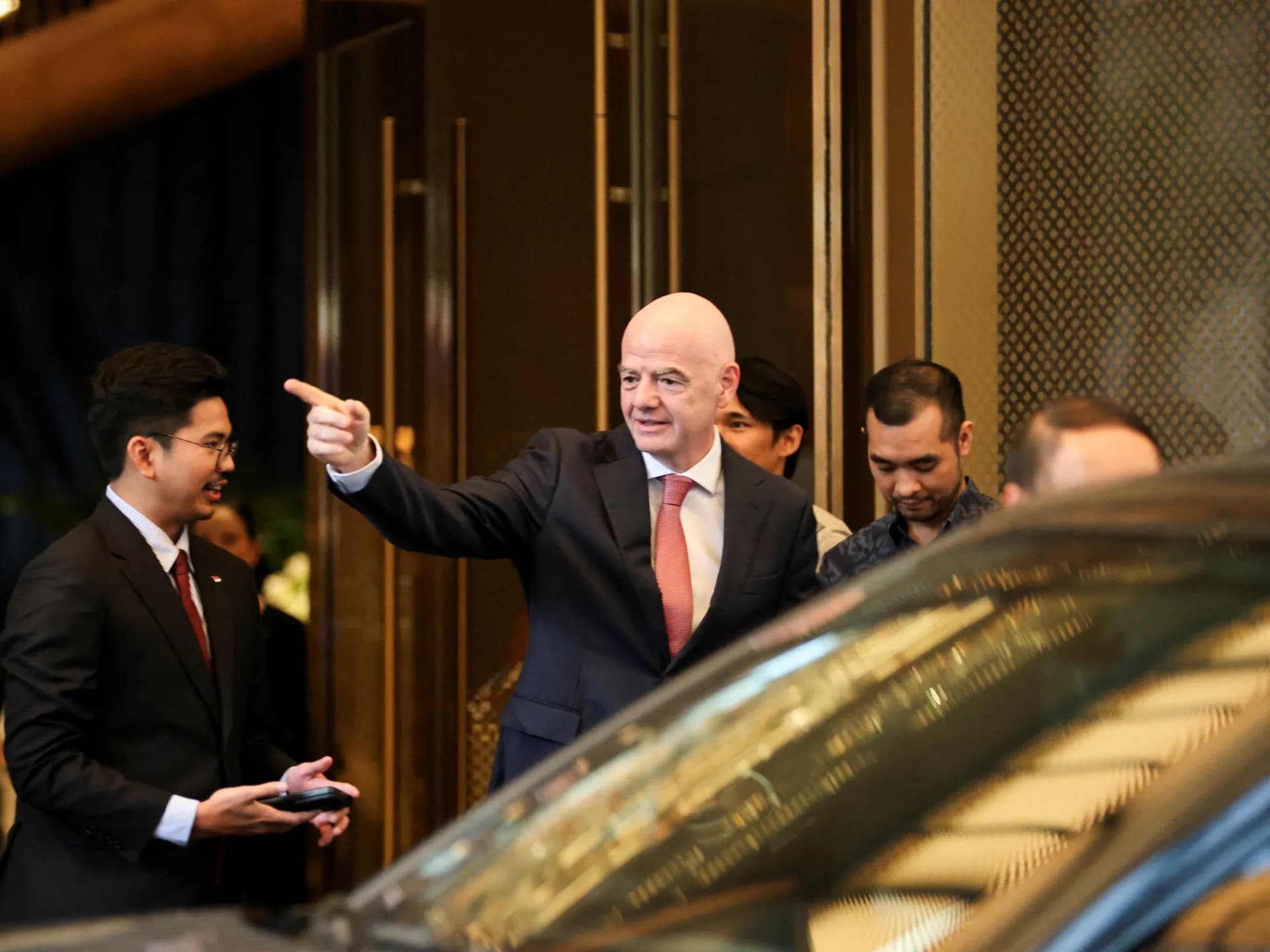 Gianni Infantino, de traje oscuro y corbata roja, señala con el brazo extendido al salir de un hotel rodeado de acompañantes y junto a un automóvil negro