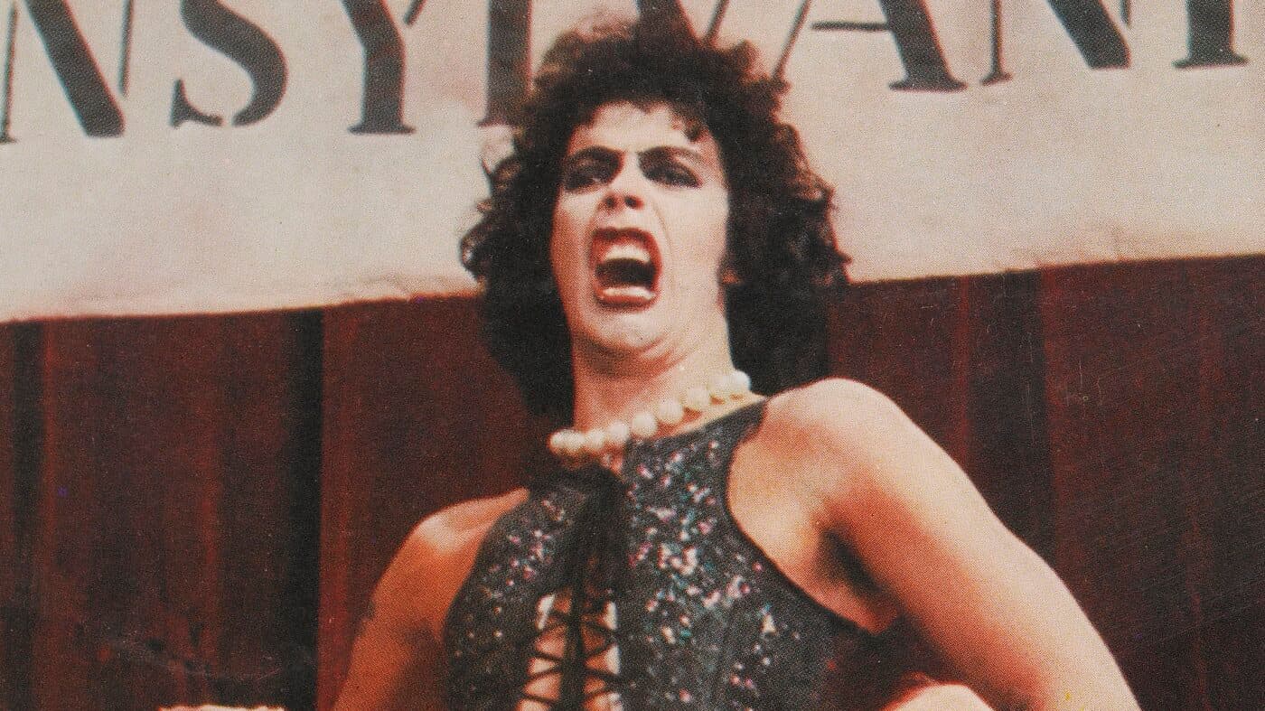 Tim Curry, caracterizado como Frank-N-Furter con maquillaje oscuro en los ojos, collar de perlas y corsé de lentejuelas, canta con la boca abierta