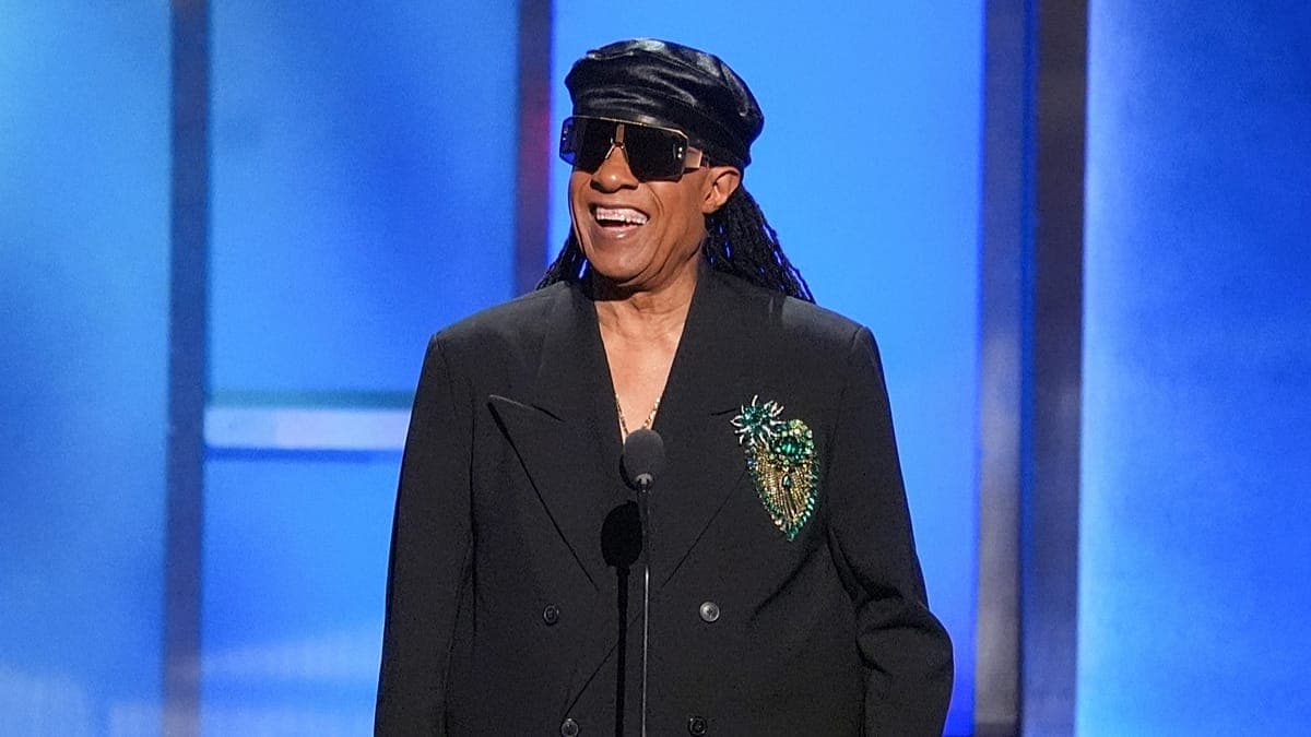 Stevie Wonder, con gafas oscuras y gorra de cuero, sonríe frente a un micrófono en un escenario